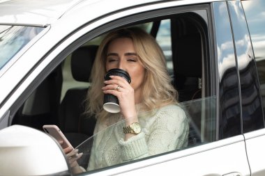 Güzel kadın oturuyor ve araba kullanıyor. Elinde bir cep telefonu ve mesajlaşma var. Bayan kahve içiyor ve mutlu bir şekilde dört gözle bekliyor. Gülümsüyor.