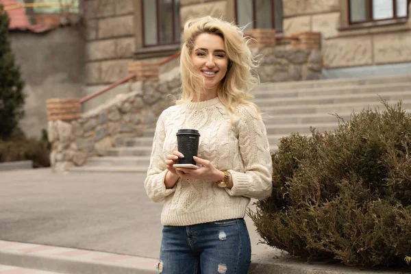 Modern young woman in a big city. Blonde woman with cell phone and cup coffee.