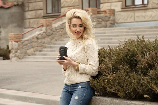 Modern young woman in a big city. Blonde woman with cell phone and cup coffee.
