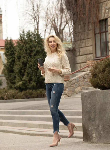 Modern young woman in a big city. Blonde woman with cell phone and cup coffee.