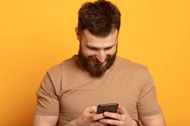 Sarı arka planda izole genç sakallı adamın yakın çekim portre, onun akıllı telefon ekranda tedirgin görünümlü gülümseyerek ve mutlu bir şekilde gülüyor, web medya içeriği etkilendim