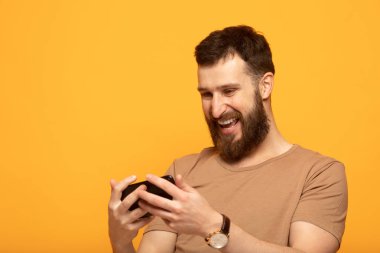 Sarı arka planda izole genç sakallı adamın yakın çekim portre, onun akıllı telefon ekranda tedirgin görünümlü gülümseyerek ve mutlu bir şekilde gülüyor, web medya içeriği etkilendim