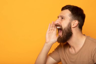 Dikkat! T-shirt genç brunet sakallı adam yakın portre metin için kopya alanı ile canlı sarı arka plan izole onu açık ağız yakınında el tutuyor