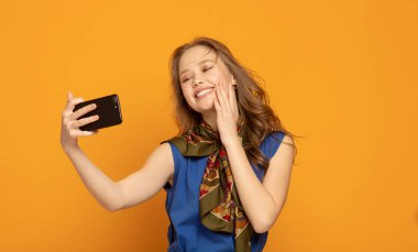 Selfie zamanı! Çekici genç kadın rahat giyim onu akıllı telefon ile selfie yapma ve sarı arka plan karşısında dururken gülümseyerek