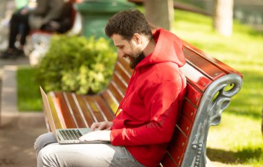 Pozitif Kafkas sakallı adam açık havada çalışan genel dizüstü bilgisayar önünde oturuyor. Açık dizüstü bilgisayar ile parkta oturan serbest çalışma