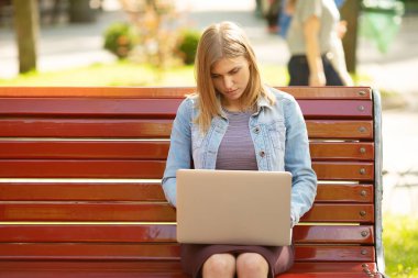 Front view of a happy student girl working with a laptop in a green park near university campus.