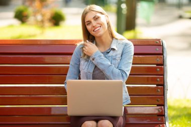Front view of a happy student girl working with a laptop in a green park near university campus.