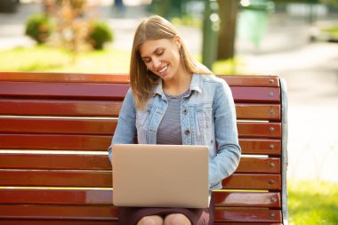 Front view of a happy student girl working with a laptop in a green park near university campus.