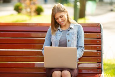 Front view of a happy student girl working with a laptop in a green park near university campus.