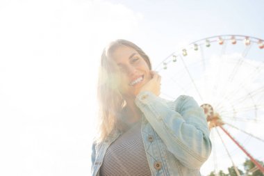 Arka planda park dönme dolap uzağa bakarak gülümseyen rahat genç sarışın yakın. Güneş ışığında mutlu kadın.