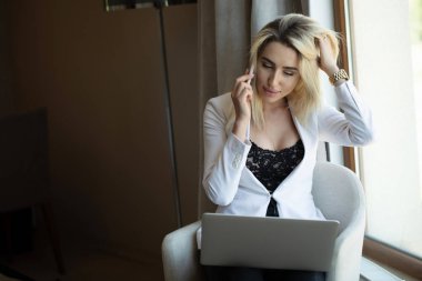 Otel odasında çalışıyorum. Takım elbiseli güzel genç iş kadını dizüstü bilgisayar üzerinde çalışıyor ve otel odasında koltukta otururken gülümsüyor.