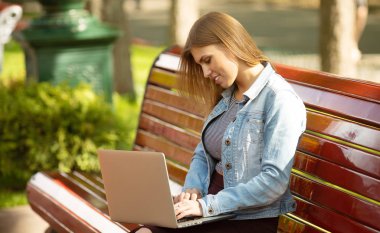Kot ceketli genç kadın parkta dizüstü bilgisayar ile çalışan bankta oturan