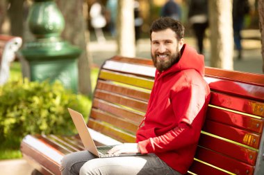 Kırmızı ceketli genç adam parkta dizüstü bilgisayar ile çalışan bankta oturan