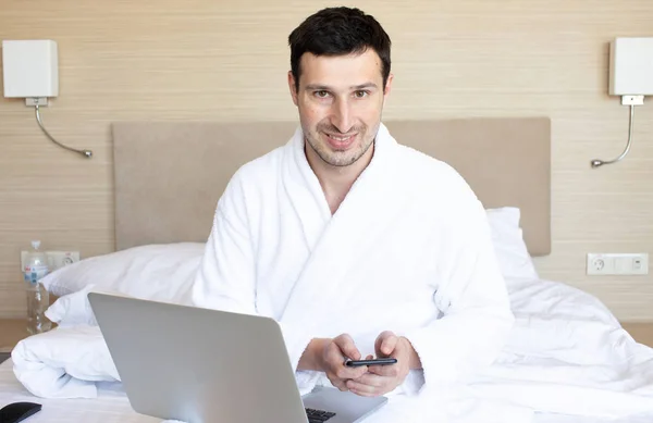 Νέος Άντρας Μπουρνούζι Χρησιμοποιώντας Φορητό Υπολογιστή Και Smartphone Στο Υπνοδωμάτιο — Φωτογραφία Αρχείου