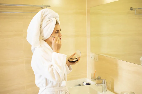 Portrait of blonde woman in bathrobe doing makeup in bathroom