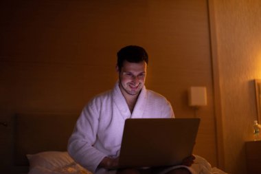 Gece yatak odasında dizüstü bilgisayar kullanarak bornoz genç adam