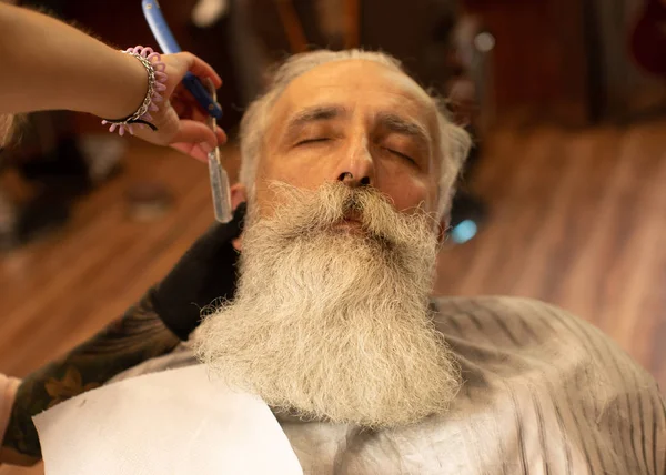 Cropped view of woman barber shaving bearded man using sharp blade.