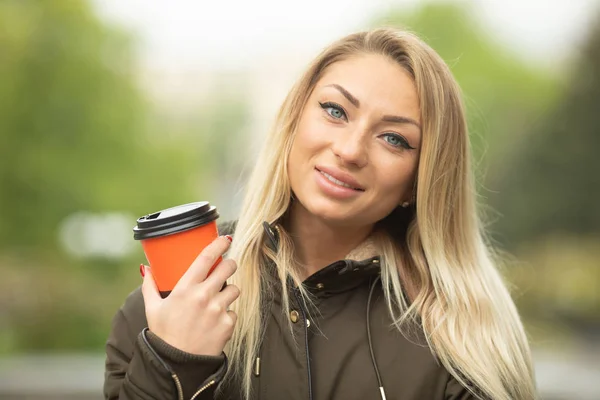 Portrait of young blonde woman in jacket drinking coffee posing on urban background