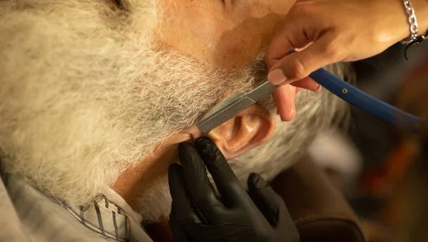 Cropped view of woman barber in black glove shaving bearded man with sharp blade.