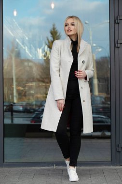 Beyaz ceketli zarif sarışın kadın şehirde poz, arka planda modern bina.