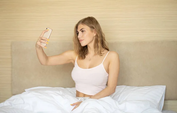 Blonde woman in pajamas using smartphone in bed