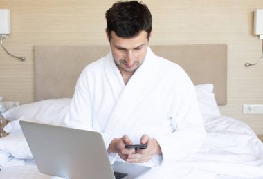 Sabah yatak odasında dizüstü bilgisayar ve akıllı telefon kullanarak bornoz genç adam