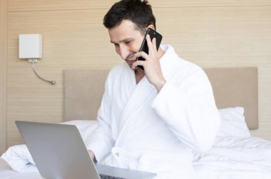 Sabah yatak odasında dizüstü bilgisayar ve akıllı telefon kullanarak bornoz genç adam