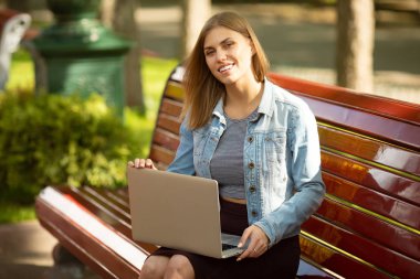 Kot ceketli genç kadın parkta dizüstü bilgisayar ile çalışan bankta oturan