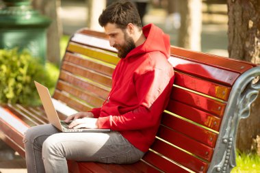 Kırmızı ceketli genç adam parkta dizüstü bilgisayar ile çalışan bankta oturan