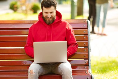 Kırmızı ceketli genç adam parkta dizüstü bilgisayar ile çalışan bankta oturan