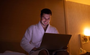 Gece yatak odasında dizüstü bilgisayar kullanarak bornoz genç adam