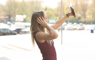 Bordo elbiseli genç sarışın kadın, kentsel arka planda poz veren akıllı telefonu kullanarak selfie çekiyor