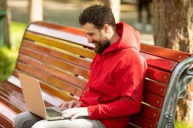 Kırmızı ceketli genç adam parkta dizüstü bilgisayar ile çalışan bankta oturan