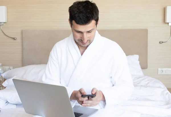 Mladý Muž Županu Který Používá Laptop Smartphone Ložnici Ráno — Stock fotografie