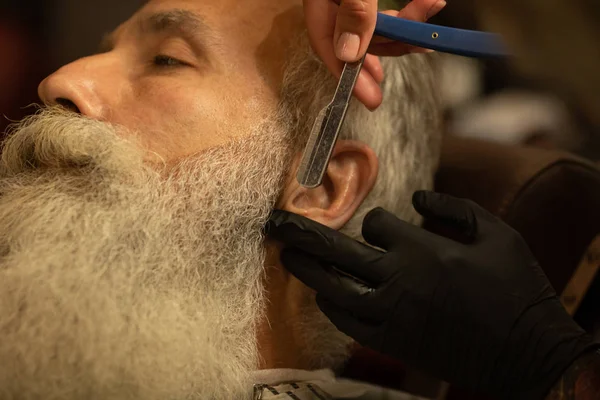 Cropped view of woman barber in black glove shaving bearded man with sharp blade.