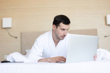 Sabah yatak odasında dizüstü bilgisayar kullanarak bornoz genç adam