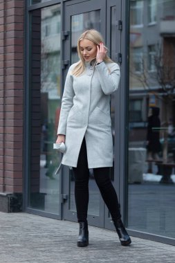 Şehirde poz veren açık gri ceketli zarif sarışın kadın, arka planda modern bina.