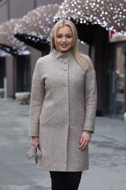 Sıcak bej yünlü ceket genç sarışın kadın şehirde poz, arka planda şenlikli ışıklar ile modern bina.