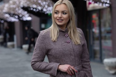 Kahverengi yünlü ceket sarışın kadın şehirde poz, arka planda şenlikli ışıklar ile modern bina.