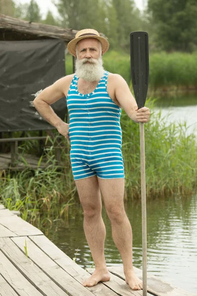 Bearded senior man in a retro swimsuit stands with a paddle on the background of the lake, paddleboard athlete.