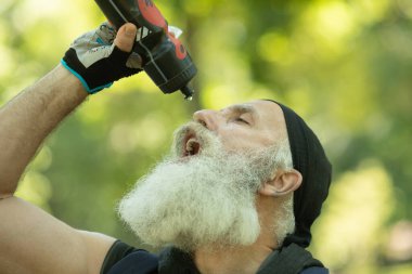 Formda olan son sınıf öğrencisi bisiklete binerken içme suyu içer. Sakallı yaşlı sporcu parkta bisiklet sürerken mola veriyor..