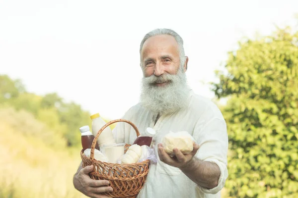 Professional bearded senior farmer is holding a basket with bottles of milk, butter, cheese. He is looking at this healthy food and smiling.