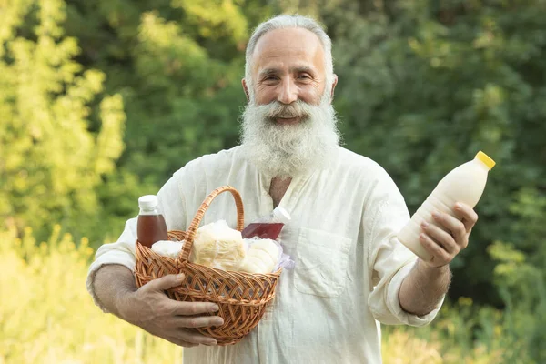 Professional bearded senior farmer is holding a basket with bottles of milk, butter, cheese. He is looking at this healthy food and smiling.
