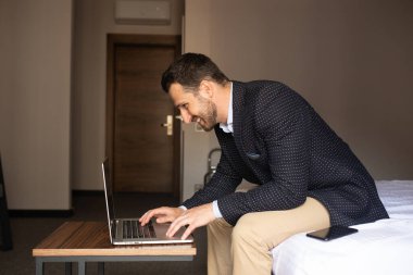 Ofisle irtibatta kalmak. Güzel takım elbiseli genç adam dizüstü bilgisayarda çalışıyor ve otel odasında yatakta otururken gülümsüyor..
