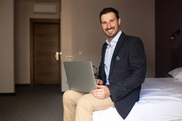 Staying in touch with office. Handsome young man in suit working on laptop and smiling while sitting on the bed in hotel room.