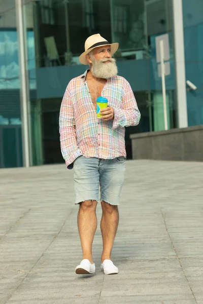 Portrait of positive senior bearded hipster man having coffee break outdoors on urban settings.