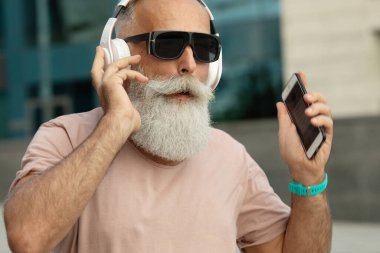 Yetişkin hippi adam şehirde yürürken kulaklıkla müzik dinleyerek eğleniyor.