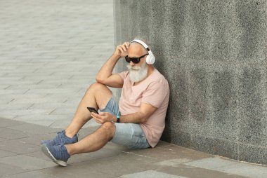 Hippi olgun bir adam şehirde sokakta oturmuş akıllı telefondan müzik dinliyor.