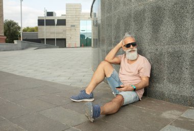 Hippi olgun bir adam akıllı telefon kullanıyor. Binaya yaslanıyor.