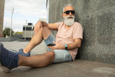 Hippi olgun bir adam akıllı telefon kullanıyor. Binaya yaslanıyor.